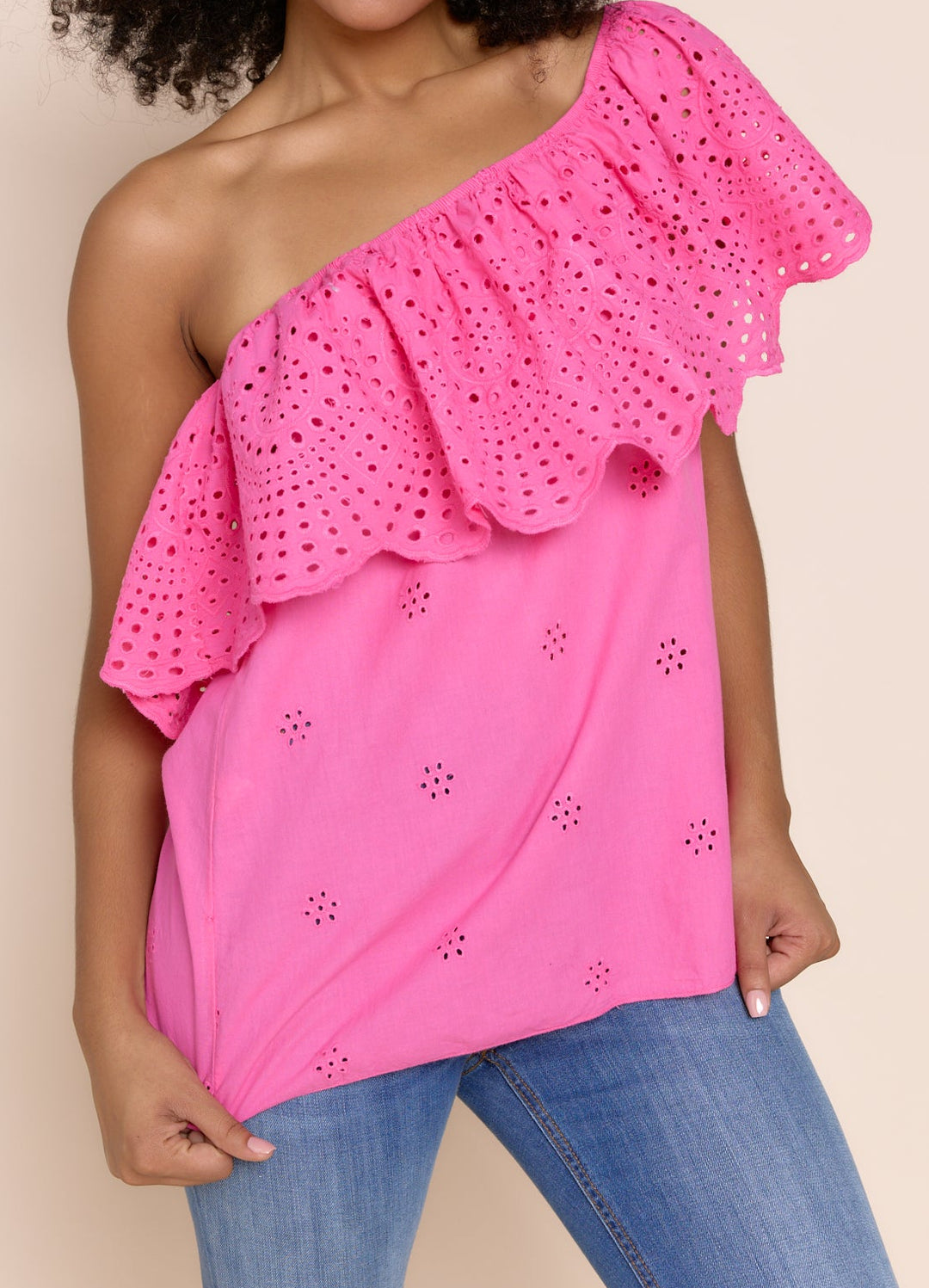 A woman modeling a bright pink one-shoulder eyelet top with a ruffled sleeve, paired with blue jeans, against a solid beige background.