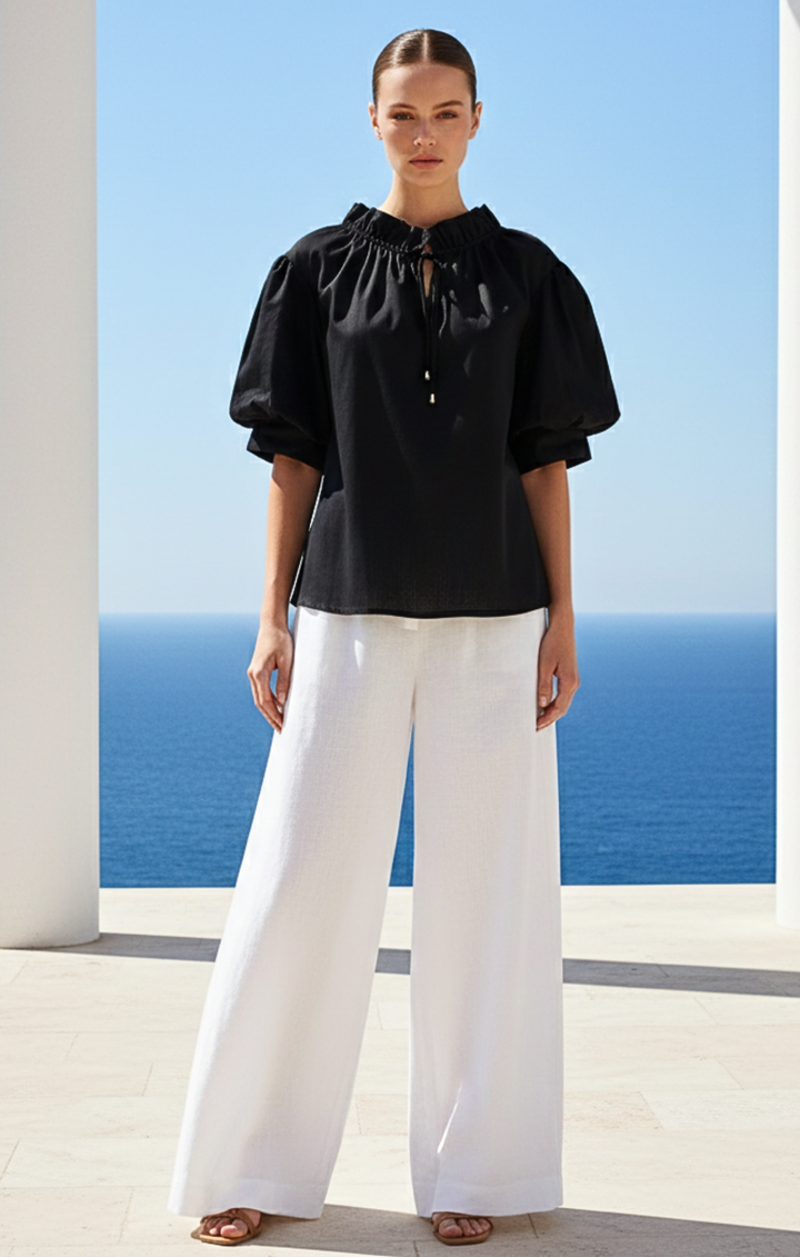 Woman wearing a black blouse and white pants standing in front of a blue ocean view.