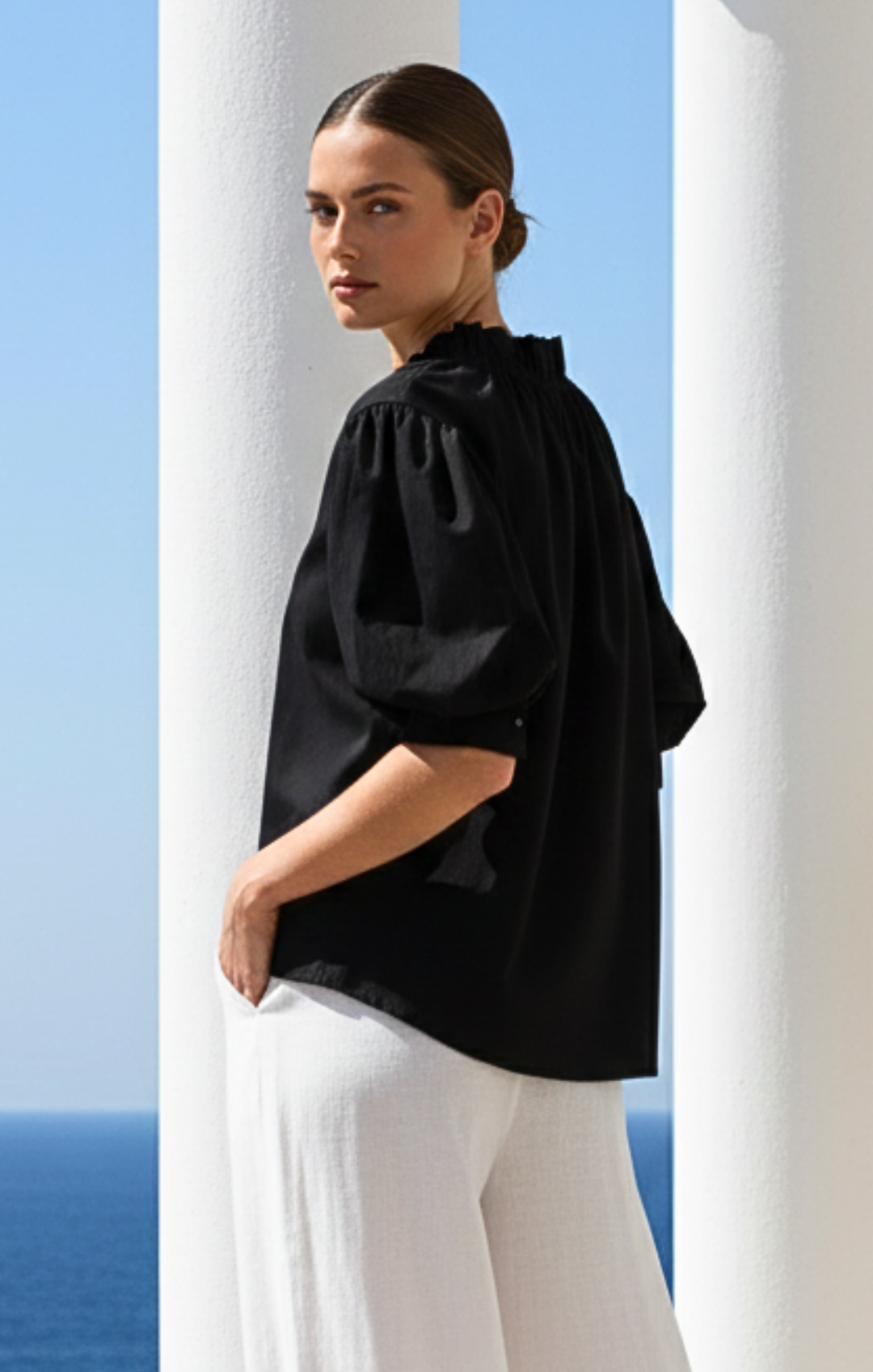 Woman wearing a black top and white pants standing against a blue sky and white columns.