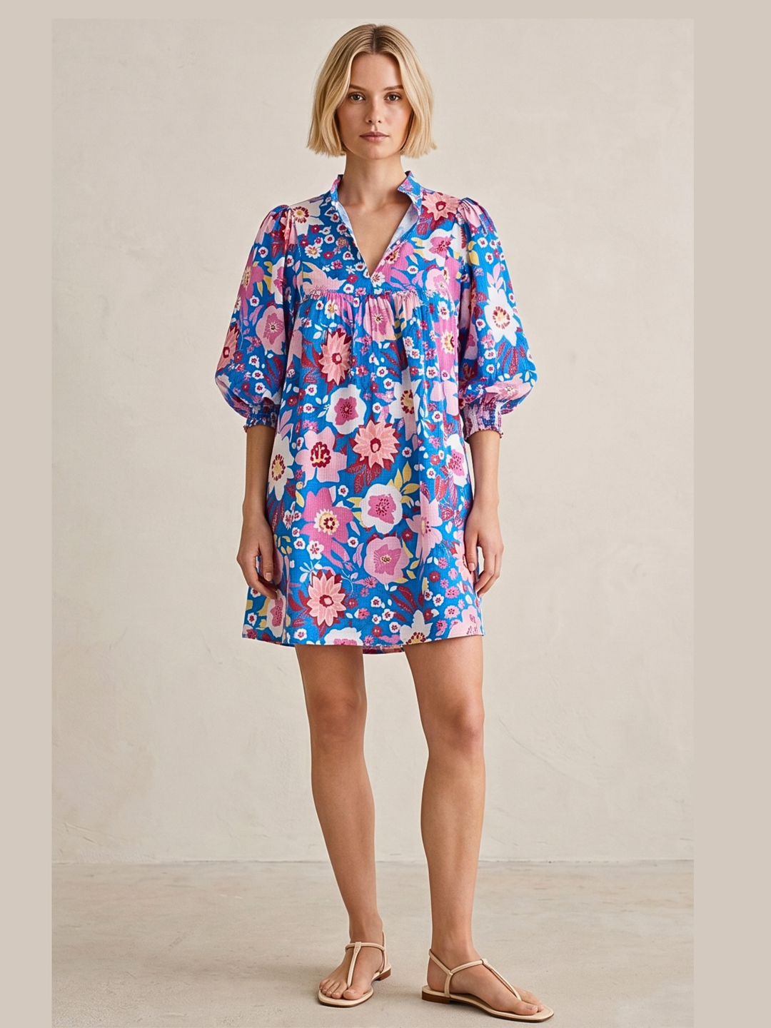 A woman modeling a short-sleeve floral dress with a blue base and pink, purple, and white flowers, paired with beige flat sandals, set against a neutral beige studio background.