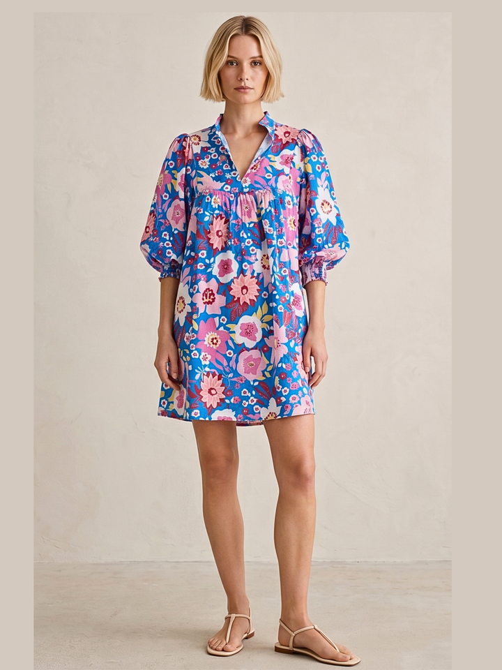 A woman modeling a short-sleeve floral dress with a blue base and pink, purple, and white flowers, paired with beige flat sandals, set against a neutral beige studio background.