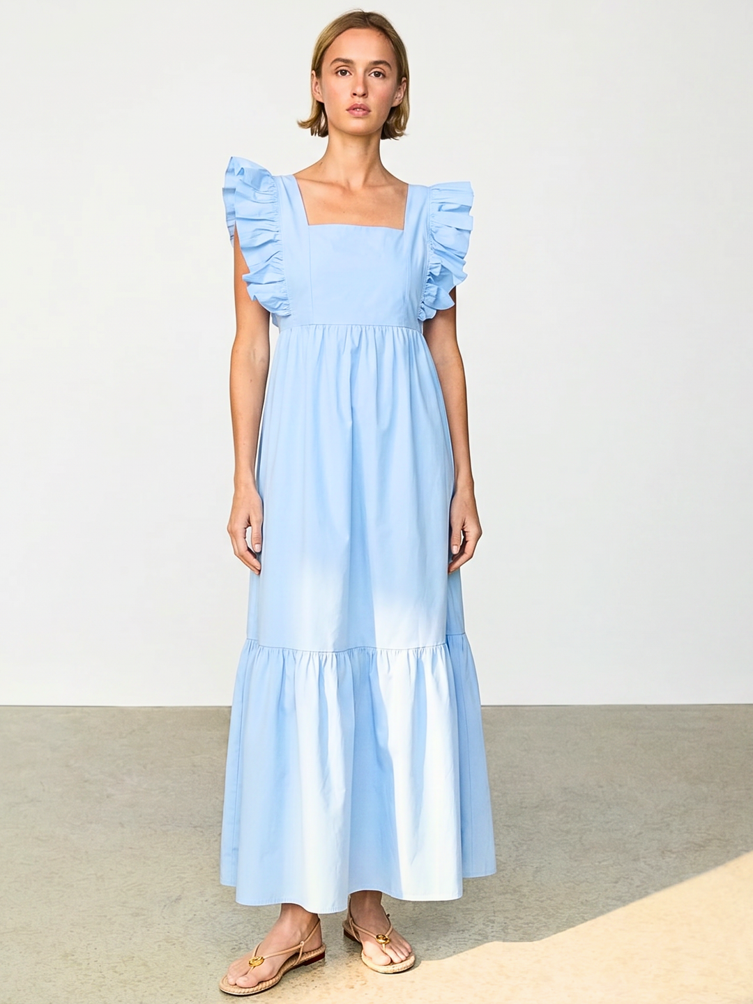 A woman modeling a light blue maxi dress with ruffled cap sleeves and a tiered skirt, standing against a plain light gray studio background.