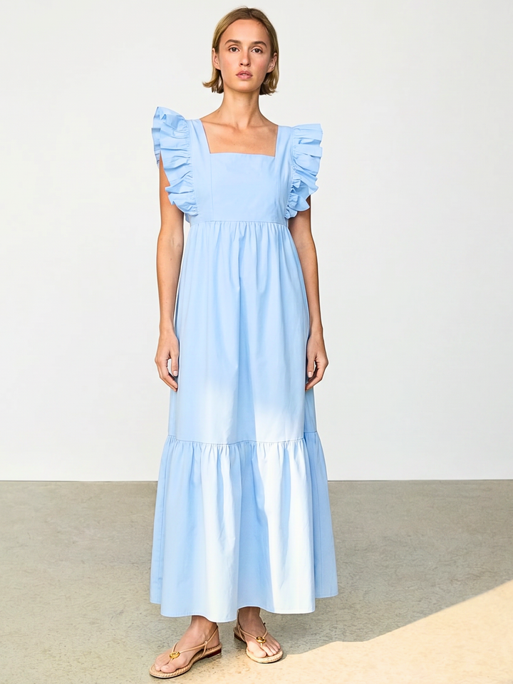 A woman modeling a light blue maxi dress with ruffled cap sleeves and a tiered skirt, standing against a plain light gray studio background.