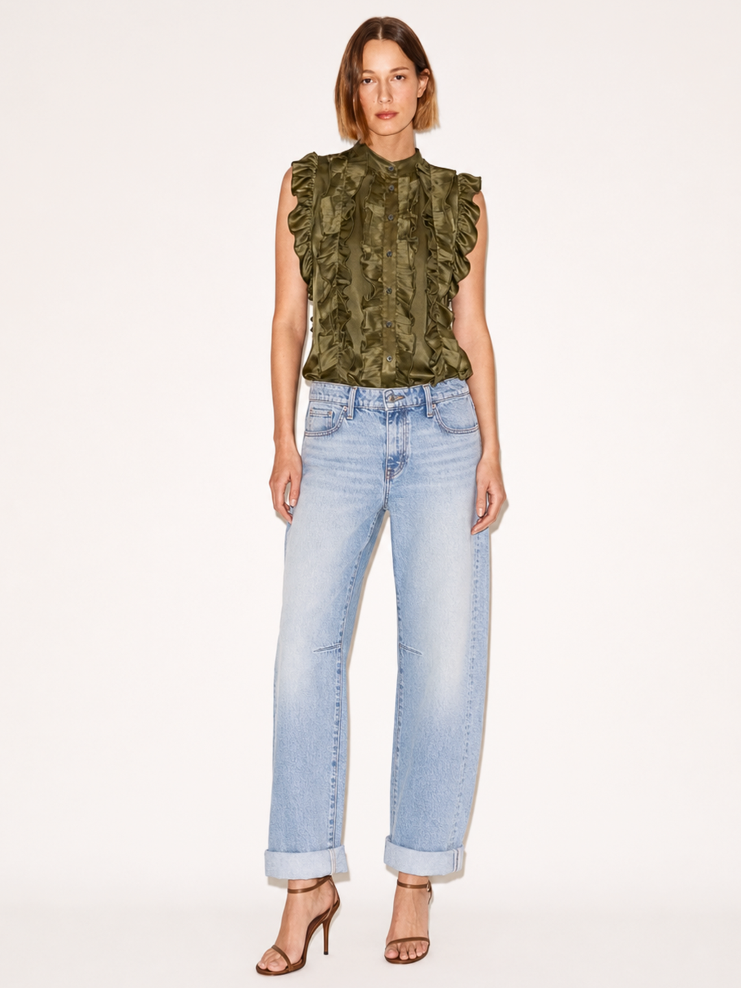 A model stands against a plain light background wearing a sleeveless olive green ruffled top, light-wash blue jeans, and brown heeled sandals.