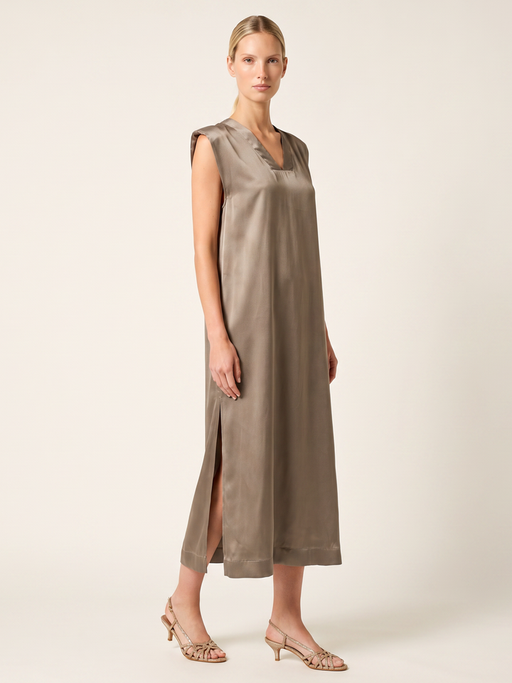 A woman modeling a sleeveless taupe midi dress with a V-neckline and side slit, paired with beige slingback heels, against a solid light beige studio background.