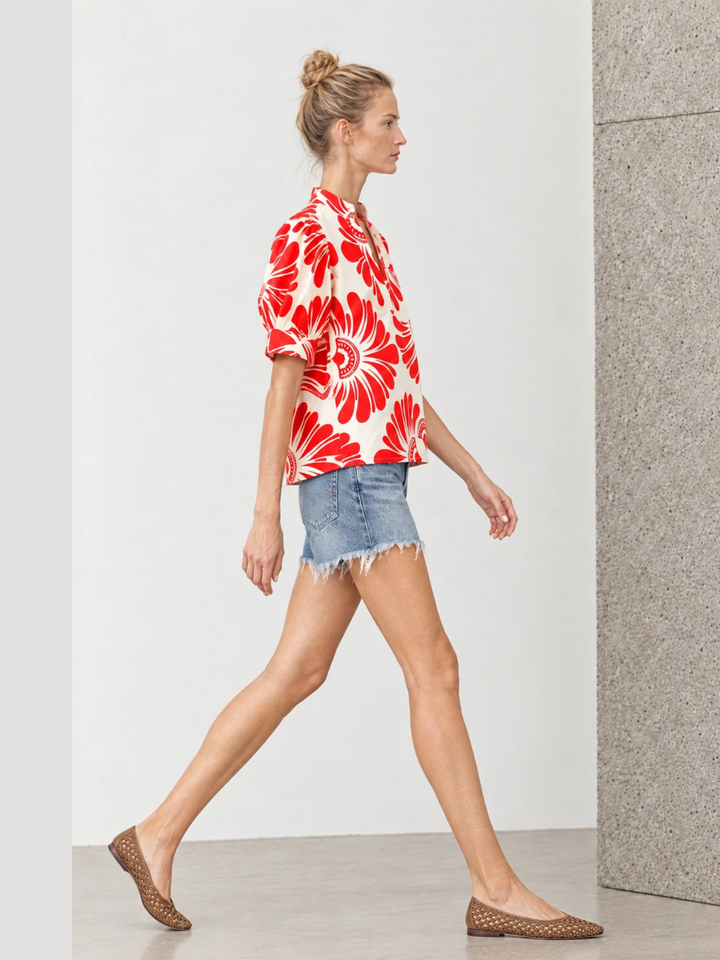 A woman walking in profile wearing a cream short-sleeve button-up shirt with a bold red floral print, denim shorts, and brown woven flat shoes in a minimalist studio setting.