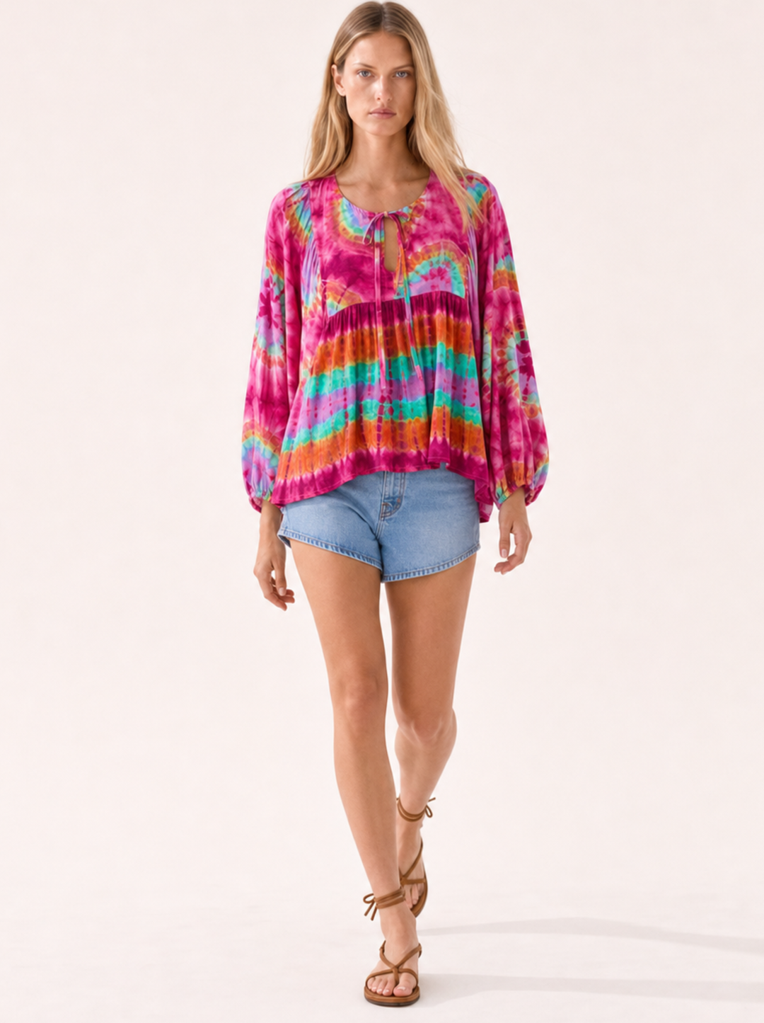A woman modeling a vibrant tie-dye long-sleeve blouse with a keyhole neckline, paired with denim shorts and brown strappy sandals, against a solid off-white background.