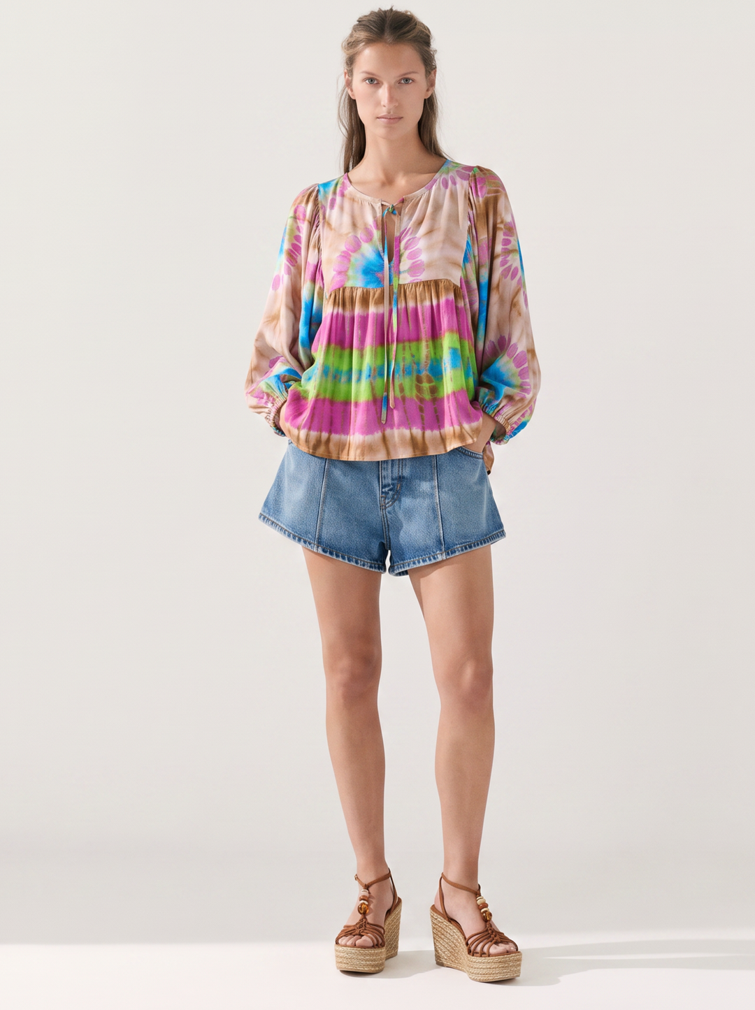 A woman modeling a long-sleeve tie-dye blouse with horizontal pastel stripes in pink, blue, green, yellow, and orange, paired with denim shorts and brown platform sandals against a light gray studio background.