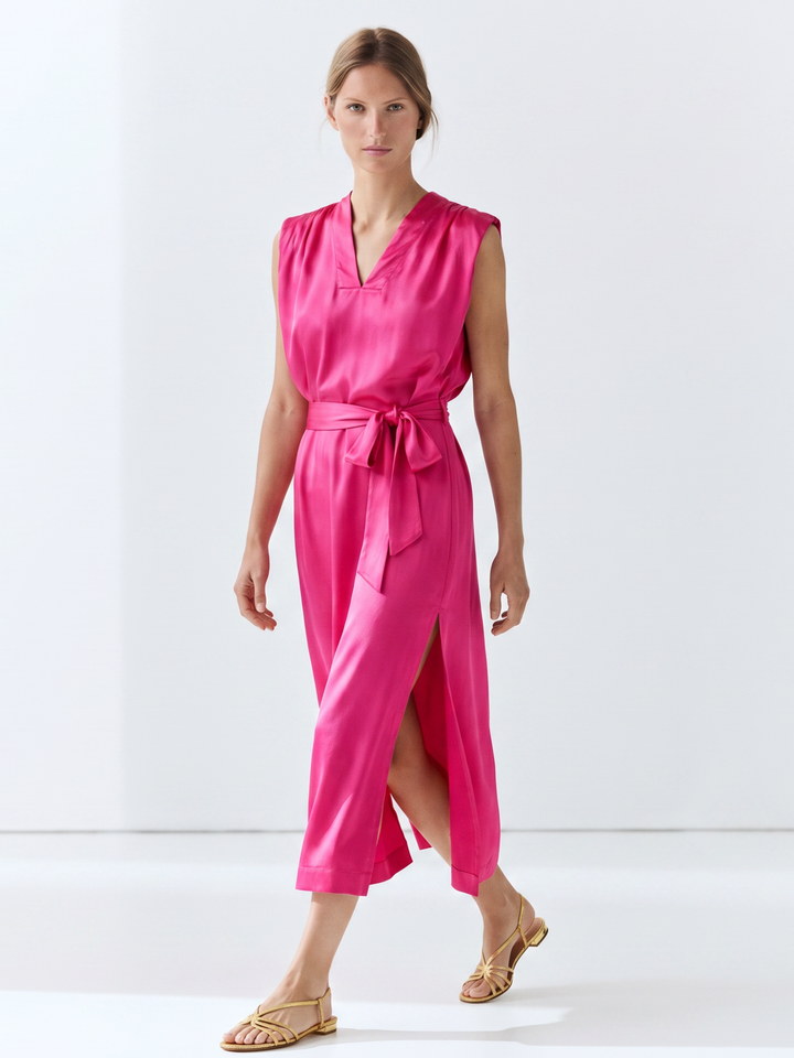 A woman modeling a bright pink satin-like midi wrap dress with a tie waist and side slit, paired with gold flat sandals, against a clean white studio background.