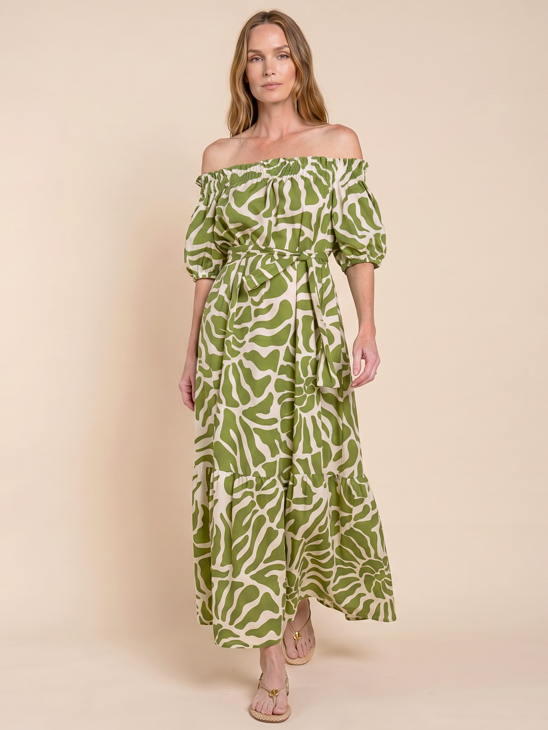 A woman modeling an off-the-shoulder maxi dress with a green and cream abstract leaf pattern, standing against a solid beige studio background.