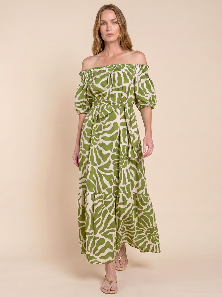 A woman modeling an off-the-shoulder maxi dress with a green and cream abstract leaf pattern, standing against a solid beige studio background.