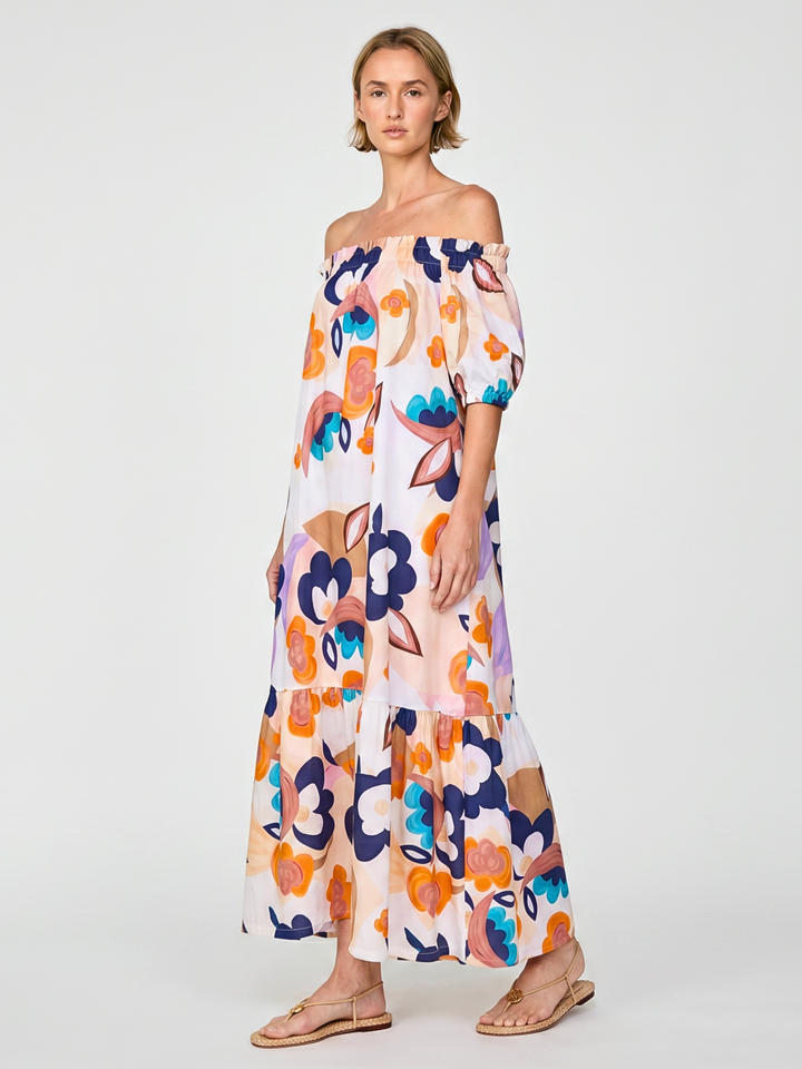 A woman modeling an off-the-shoulder maxi dress with a colorful abstract floral print on a white background, paired with flat sandals, against a light gray studio backdrop.