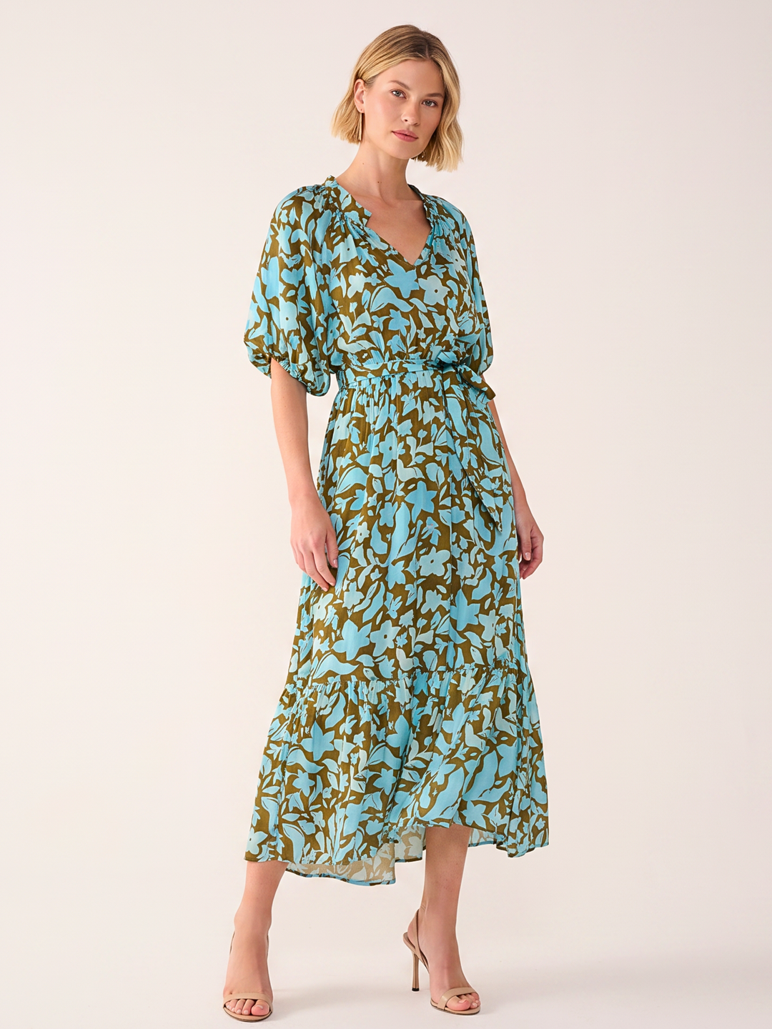 A woman modeling a light blue midi dress with an olive green abstract leaf pattern, featuring puff sleeves and a tie waist, set against a plain light beige background.