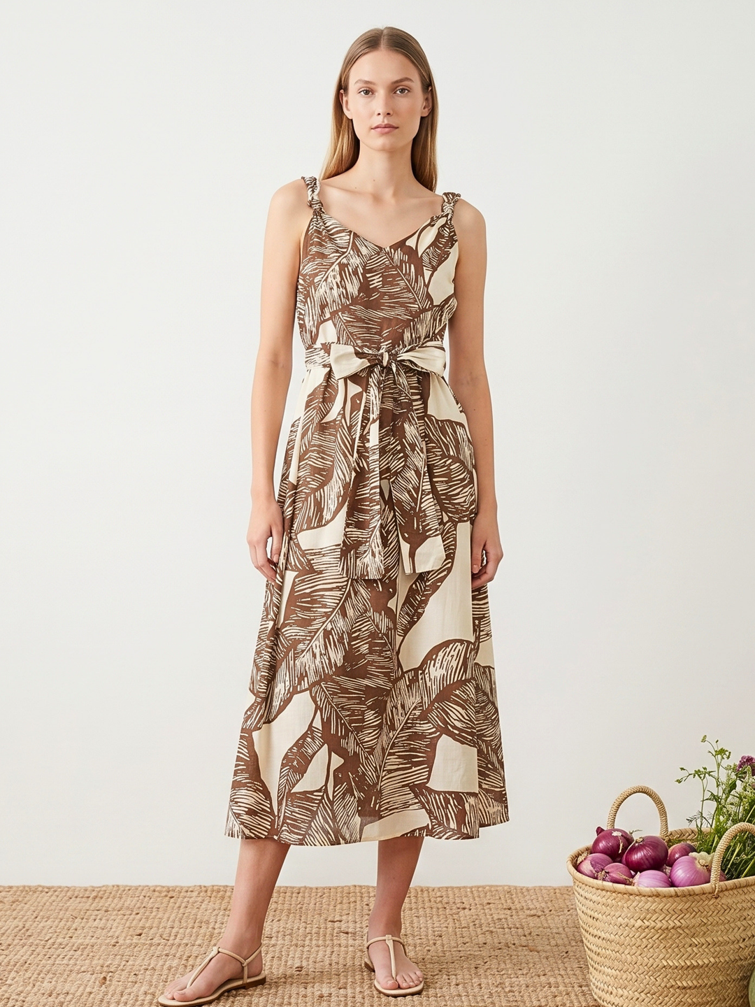 Woman wearing a leaf-patterned dress standing next to a basket of flowers and fruit on a white background