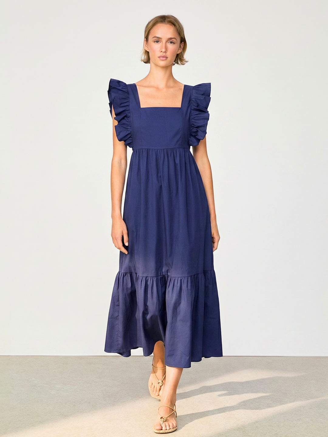 A woman modeling a navy blue midi dress with ruffled shoulder details, a square neckline, and a tiered skirt, set against a plain light gray studio background.