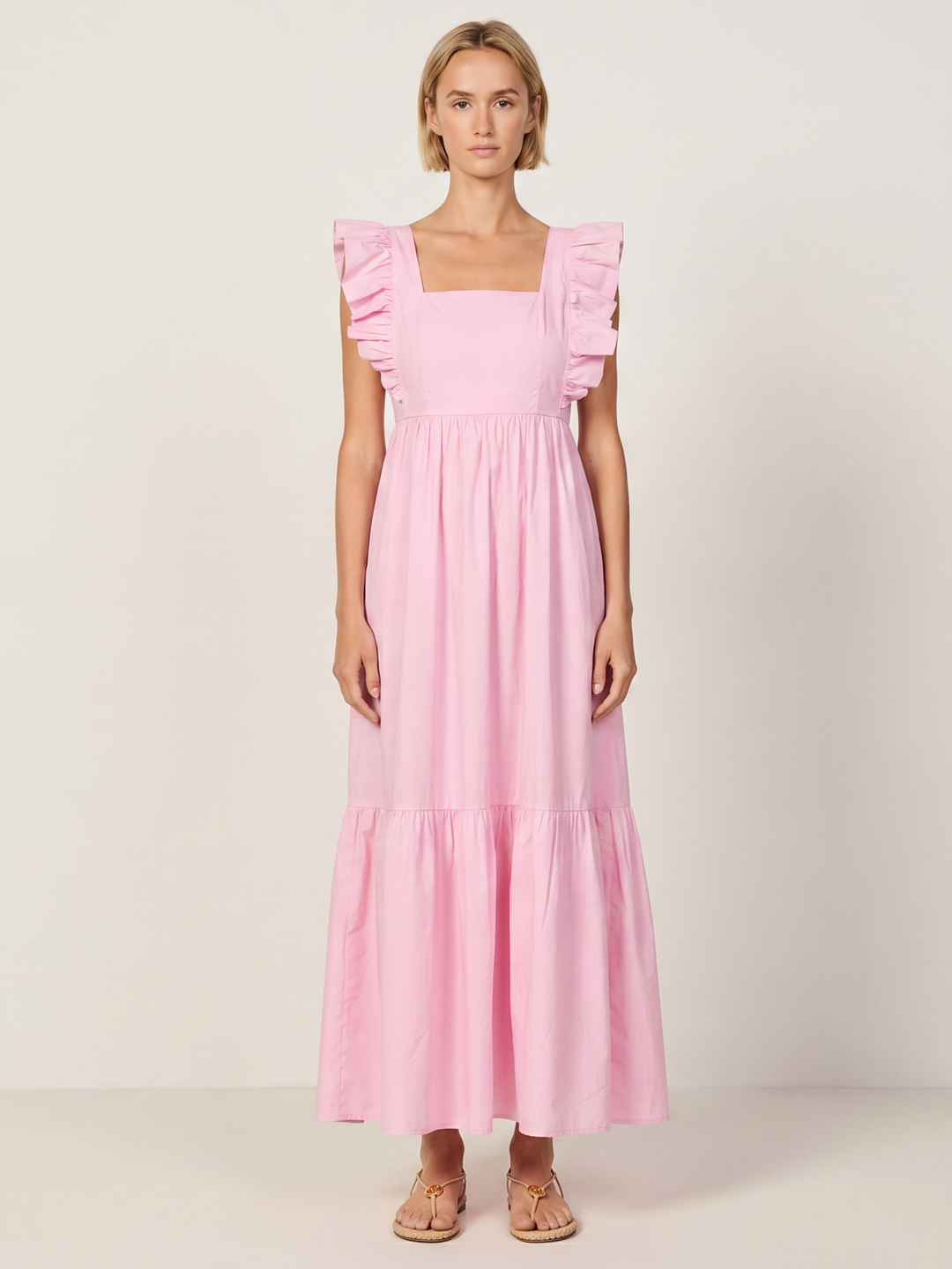 A woman modeling a light pink tiered maxi dress with ruffled cap sleeves and a square neckline, standing against a plain off-white studio background.