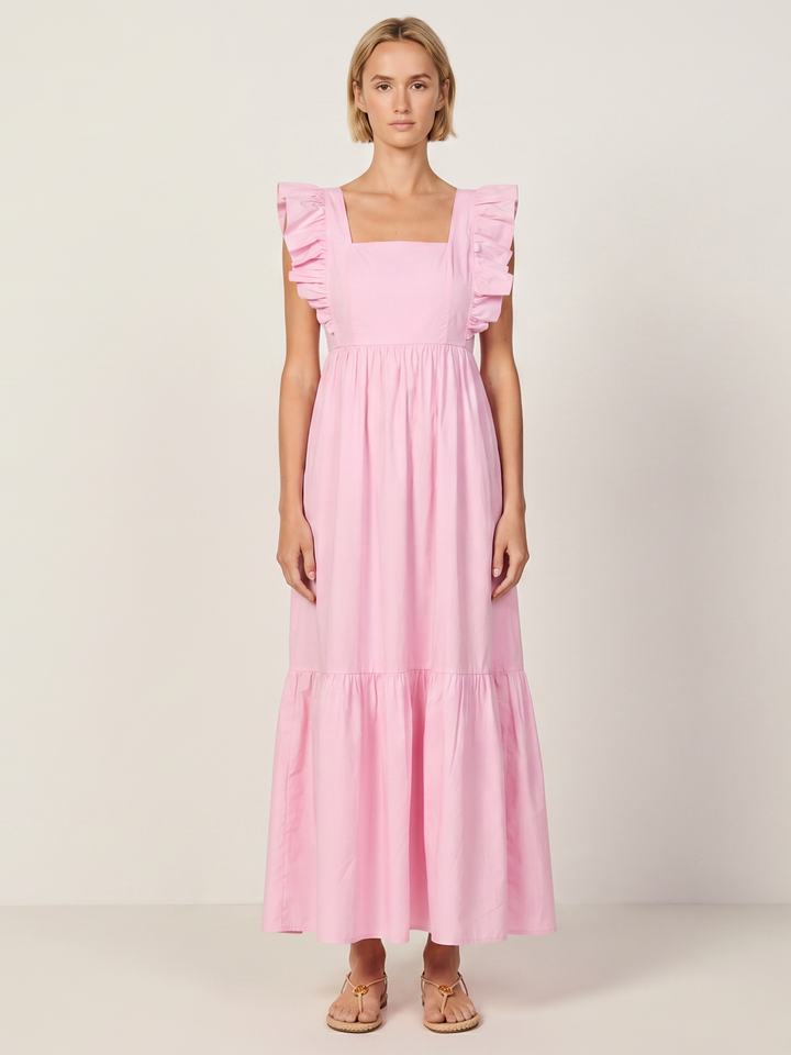 A woman modeling a light pink tiered maxi dress with ruffled cap sleeves and a square neckline, standing against a plain off-white studio background.