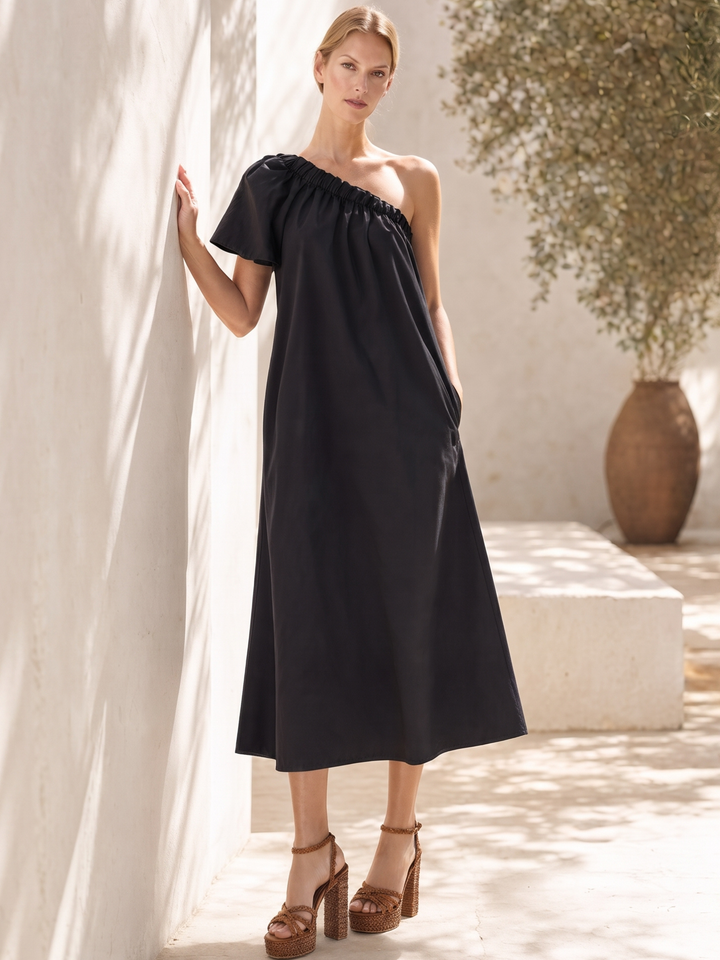 A woman models a black one-shoulder midi dress with a gathered neckline and short sleeves, standing in a sunlit outdoor courtyard next to a white wall.