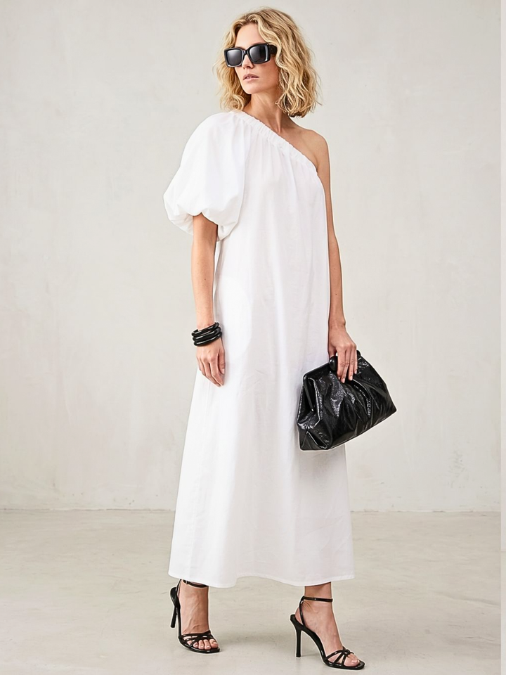 Woman wearing a white one-shoulder dress holding a black clutch against a plain background