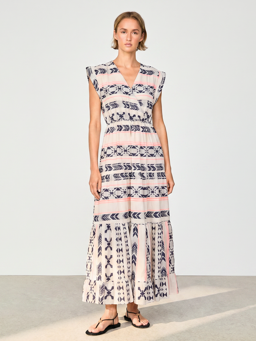 A woman modeling a sleeveless, tiered maxi dress with a pink, white, and navy blue geometric pattern, paired with black flip-flop sandals, set against a plain light gray studio background.
