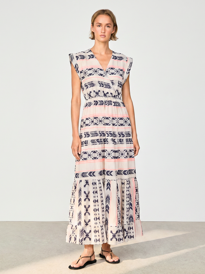 A woman modeling a sleeveless, tiered maxi dress with a pink, white, and navy blue geometric pattern, paired with black flip-flop sandals, set against a plain light gray studio background.