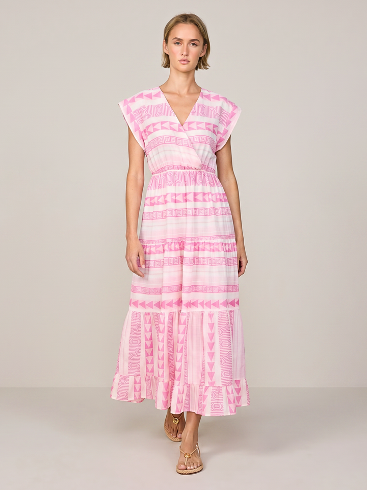 A woman modeling a pink and white patterned tiered maxi dress with a V-neckline and cap sleeves, standing against a neutral light gray studio background.