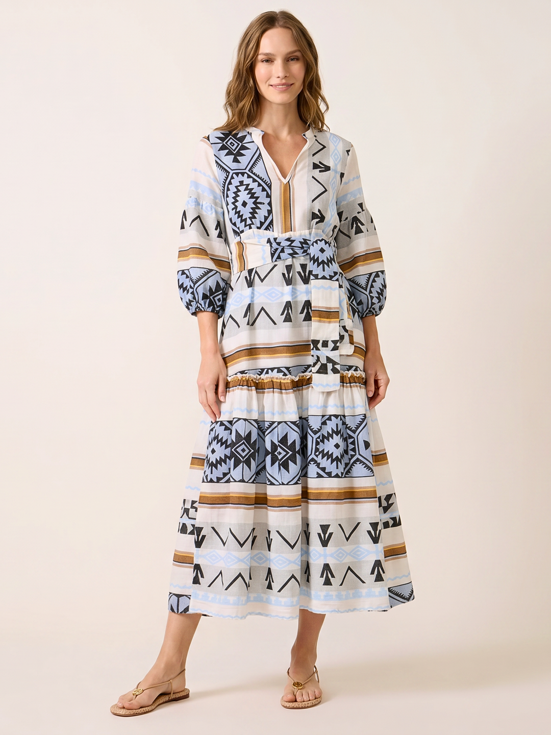 A woman modeling a bohemian-style tiered midi dress with a geometric and tribal pattern in navy blue, black, tan, and white, paired with tan flat sandals against a solid beige studio background.