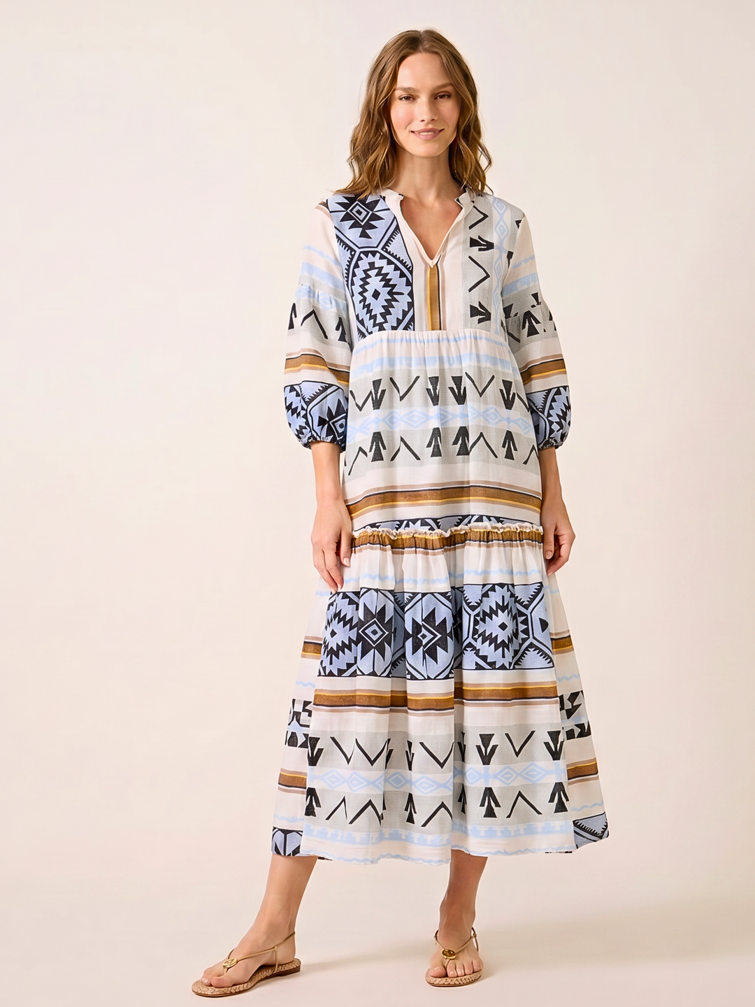 A woman modeling a tiered bohemian maxi dress with a geometric tribal pattern in navy blue, black, beige, and white, paired with tan flat sandals against a solid light beige background.