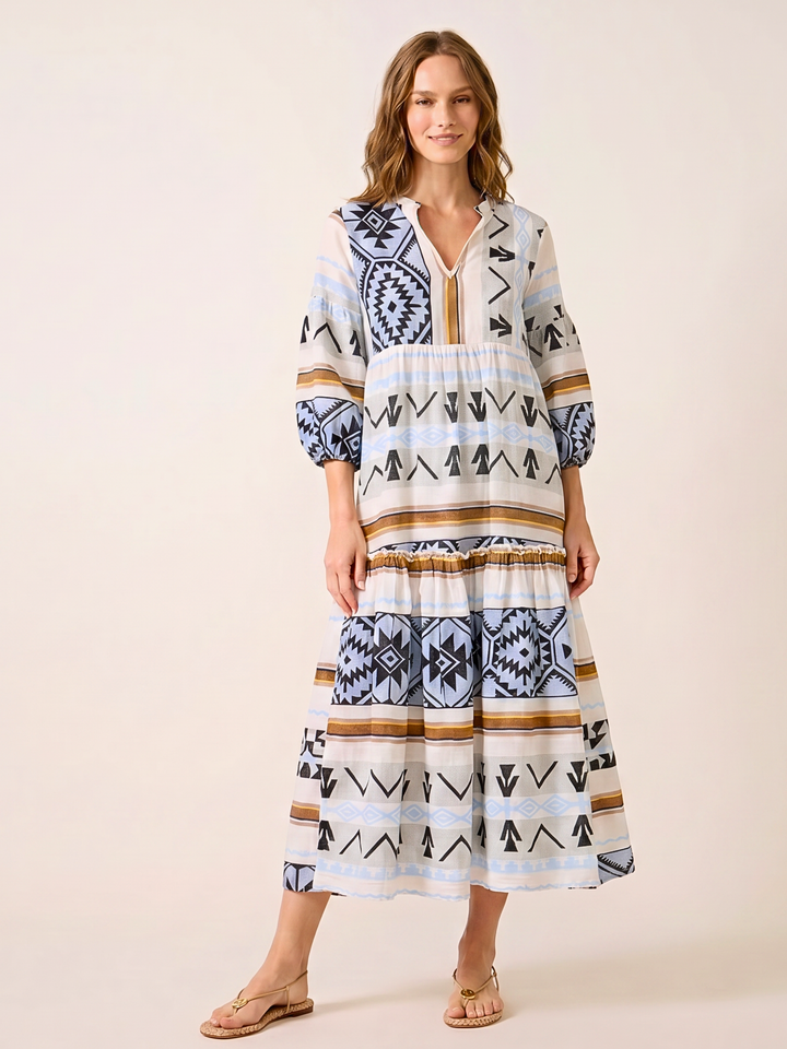 A woman modeling a tiered bohemian maxi dress with a geometric tribal pattern in navy blue, black, beige, and white, paired with tan flat sandals against a solid light beige background.