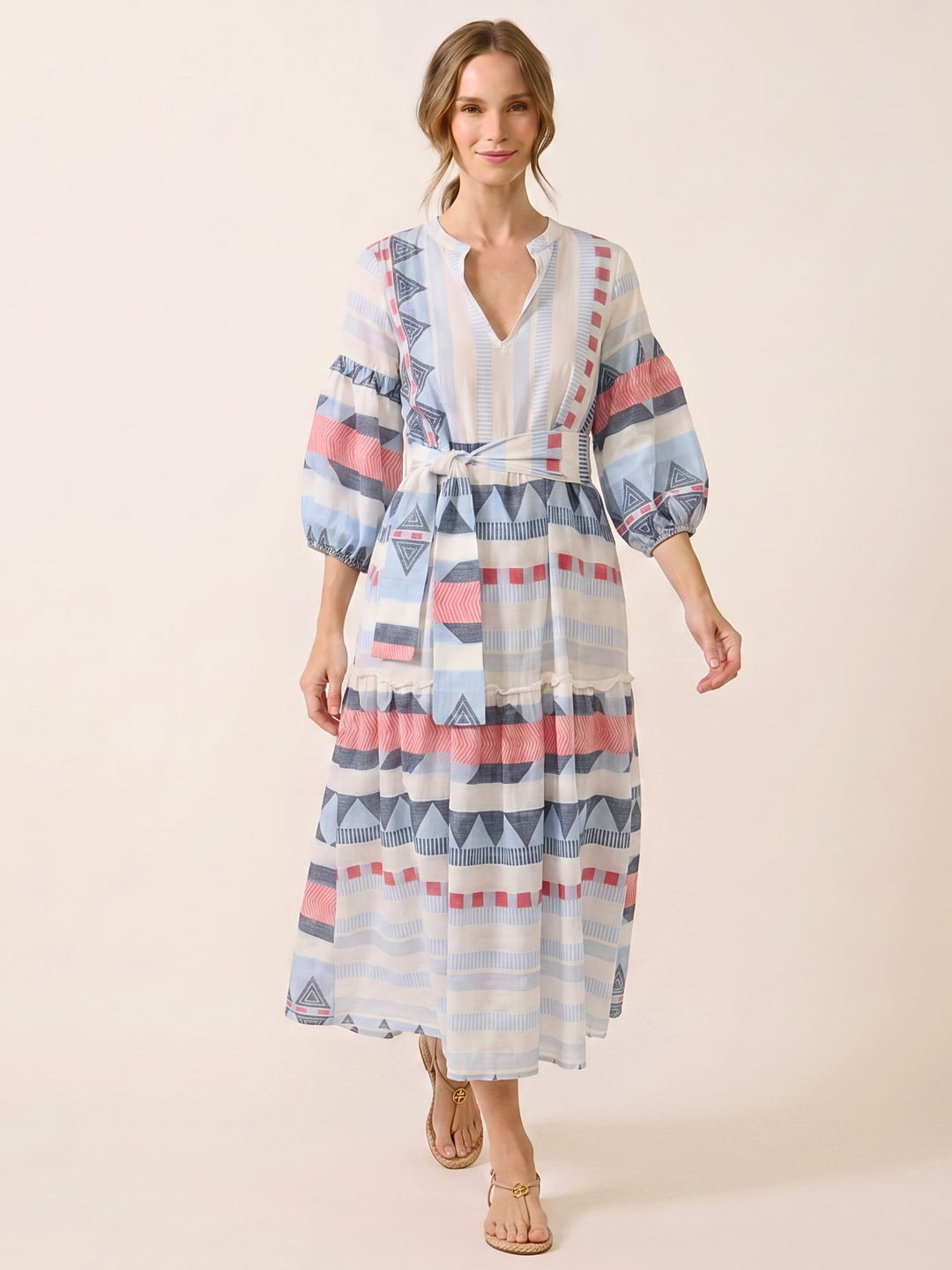 A woman modeling a tiered maxi dress with a geometric pattern in shades of blue, pink, and white, featuring balloon sleeves and a tie waist, set against a solid beige background.