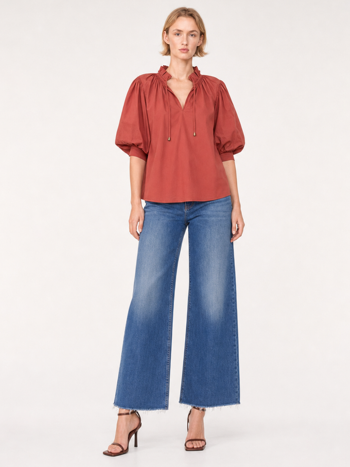 A woman modeling a rust-colored puff-sleeve blouse with a tie-neck detail, paired with wide-leg blue jeans and brown strappy heels, against a plain light gray studio background.