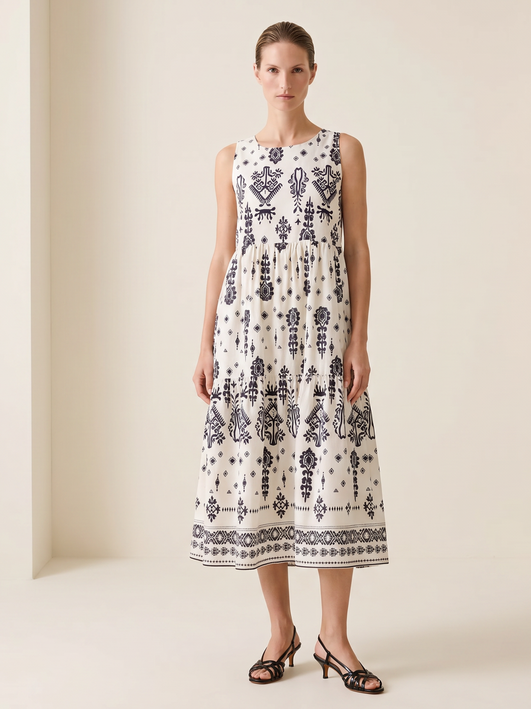 A woman stands in a studio wearing a sleeveless midi dress with a white base and dark navy blue bohemian-style geometric and floral patterns. She is wearing black strappy heeled sandals against a warm beige background.