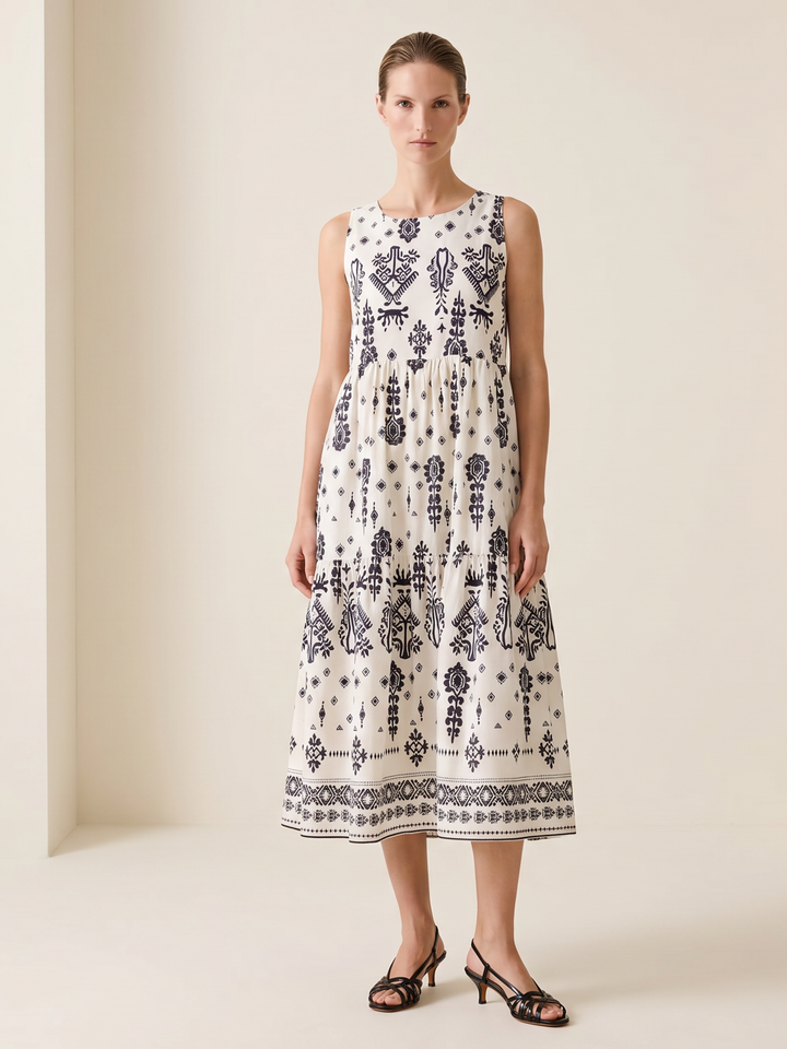 A woman stands in a studio wearing a sleeveless midi dress with a white base and dark navy blue bohemian-style geometric and floral patterns. She is wearing black strappy heeled sandals against a warm beige background.