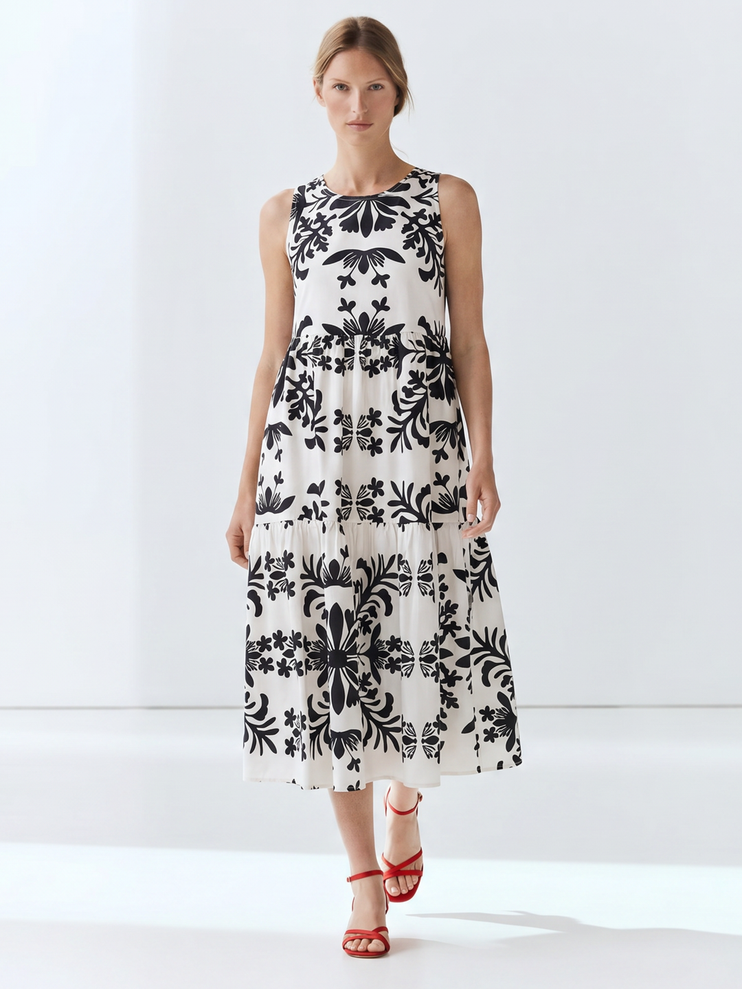 A woman modeling a sleeveless midi dress with a black floral damask pattern on an off-white background, paired with red strappy high-heeled sandals, set against a clean white studio backdrop.