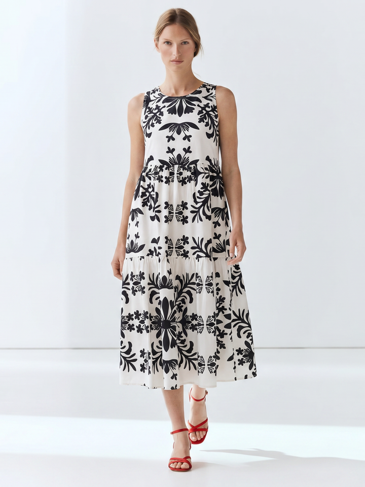 A woman modeling a sleeveless midi dress with a black floral damask pattern on an off-white background, paired with red strappy high-heeled sandals, set against a clean white studio backdrop.