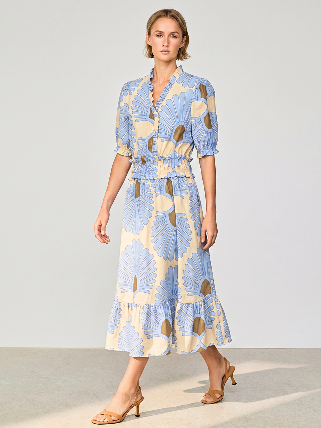 A woman modeling a light blue and cream floral print midi dress with puff sleeves and a ruffled hem, paired with tan heeled sandals against a neutral studio background.