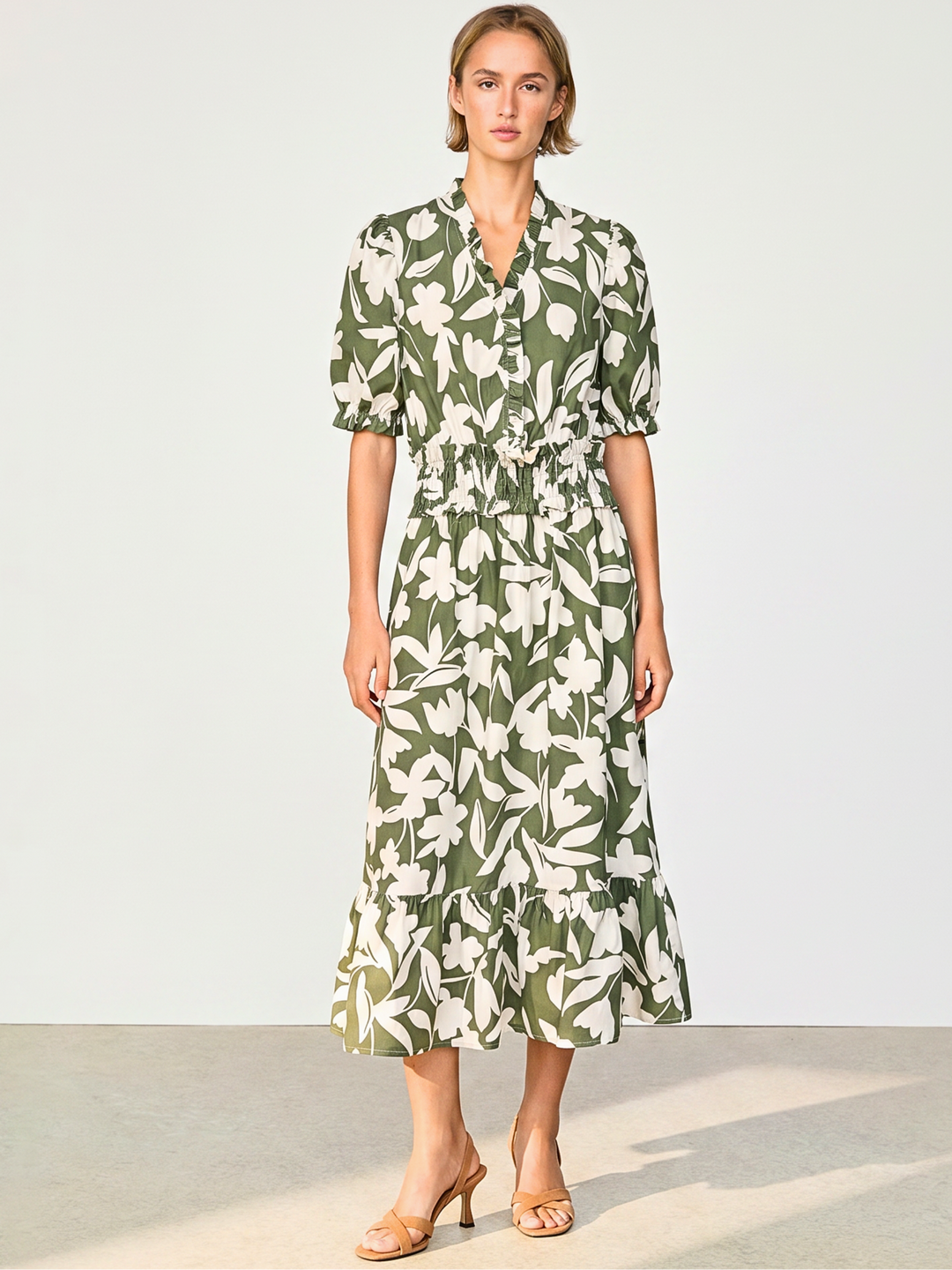 A woman modeling a green and white floral print midi dress with short puff sleeves, a button-up front, and a ruffled hem, standing against a plain light gray studio background.