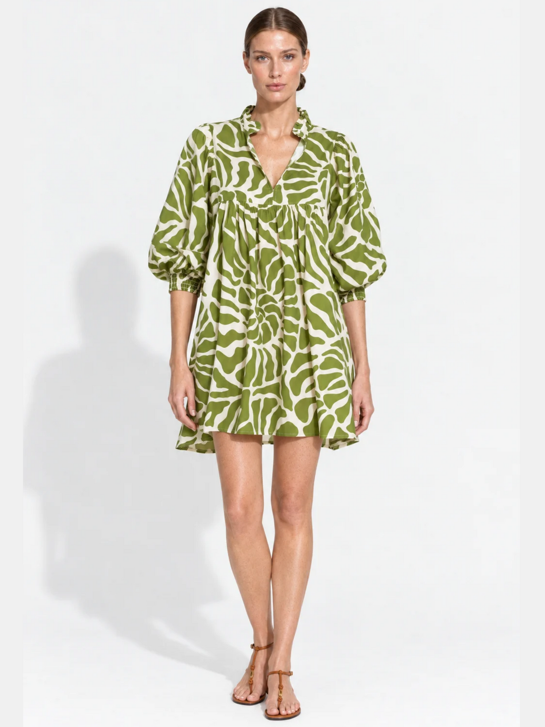 A woman modeling a short green and white abstract print dress with a V-neckline, gathered waist, and voluminous elbow-length sleeves, photographed against a plain white studio background.
