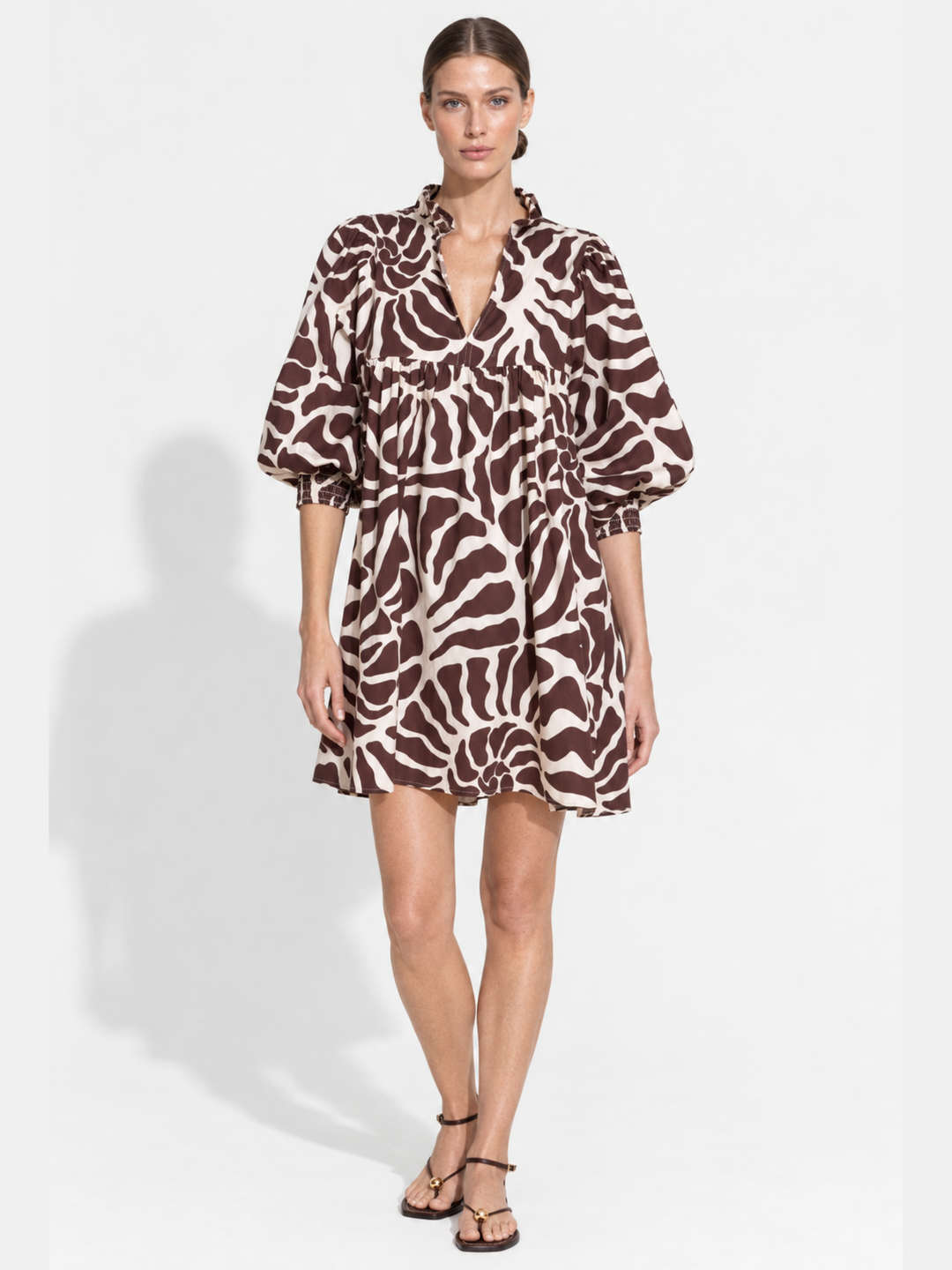 A woman modeling a brown and beige zebra-print mini dress with puff sleeves and a V-neckline, paired with black strappy sandals against a white studio background.
