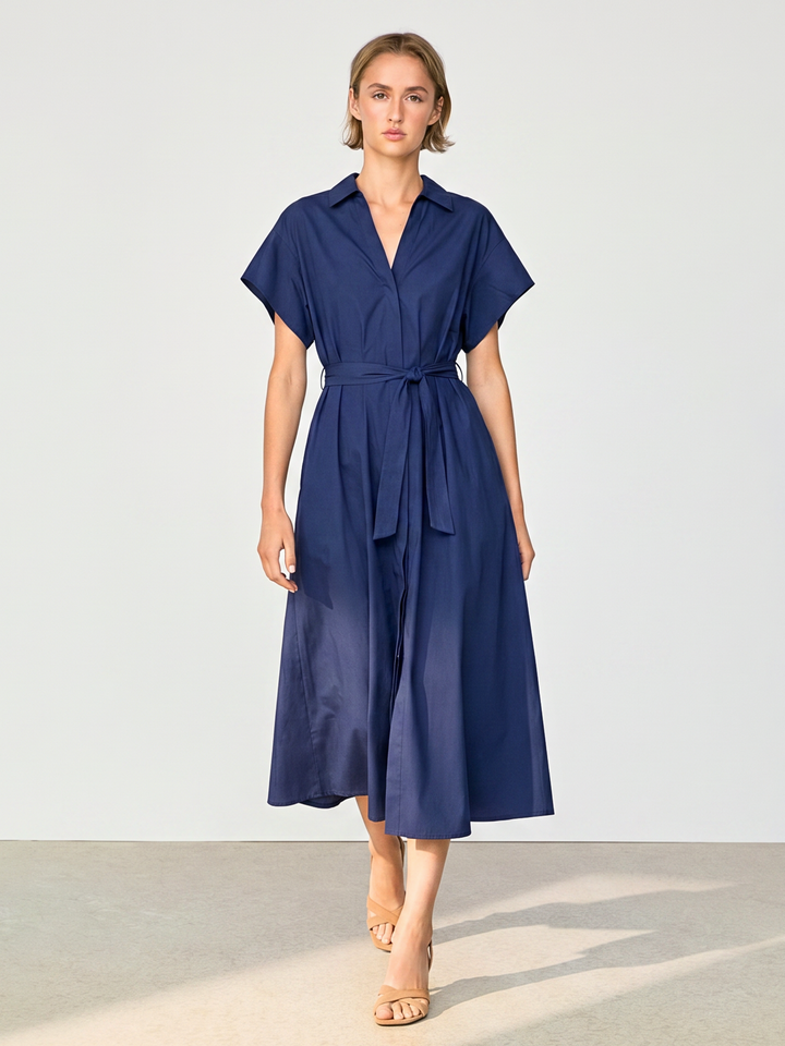 A woman modeling a navy blue midi shirt dress with short sleeves, a collared neckline, and a matching waist tie, set against a neutral studio background.