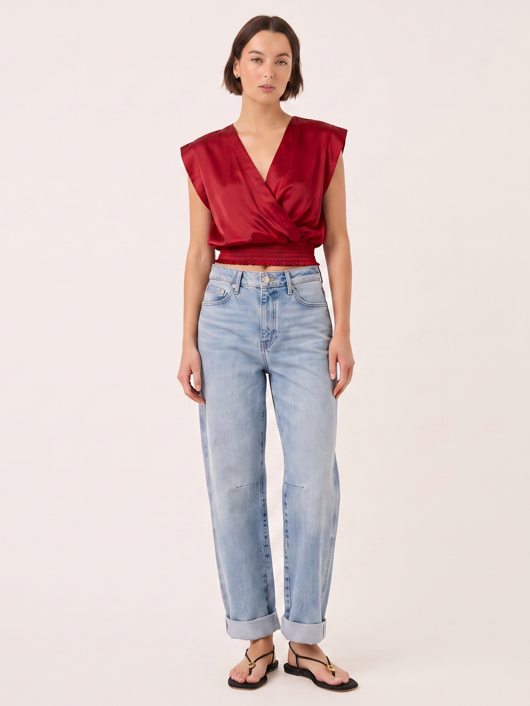 A model wearing a deep red satin wrap-style sleeveless top, light-wash blue jeans, and black sandals, standing against a solid off-white background.
