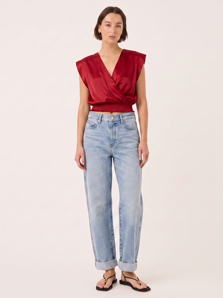 A model wearing a deep red satin wrap-style sleeveless top, light-wash blue jeans, and black sandals, standing against a solid off-white background.