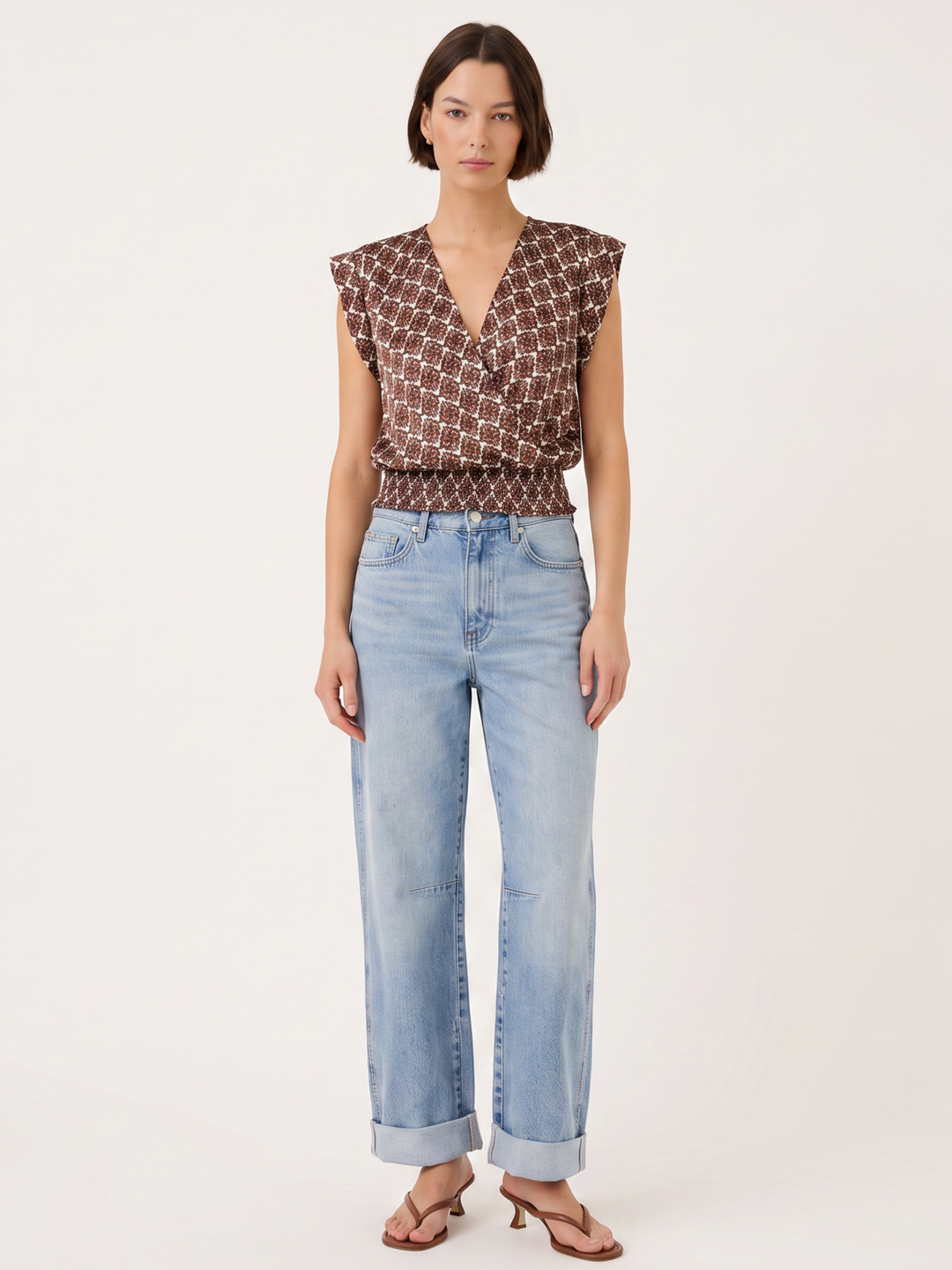 A model wearing a brown and beige geometric patterned sleeveless top with light blue wide-leg jeans and brown sandals, standing against a plain off-white studio background.
