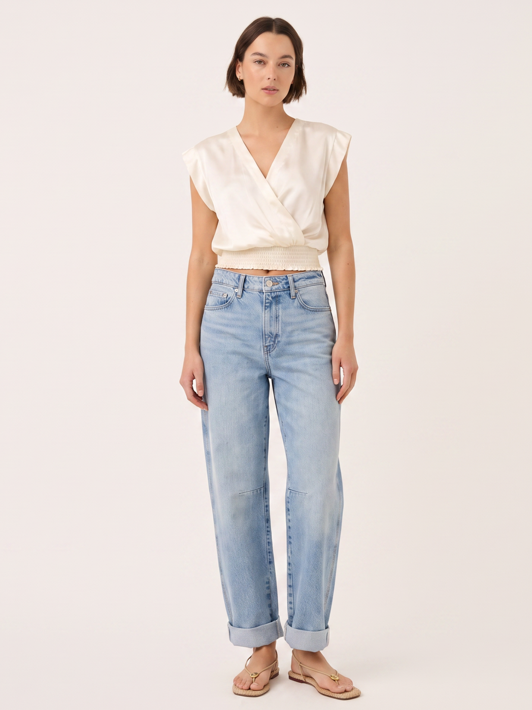 A model wearing a cream-colored sleeveless wrap top and light-wash blue jeans, standing against a solid light beige background.