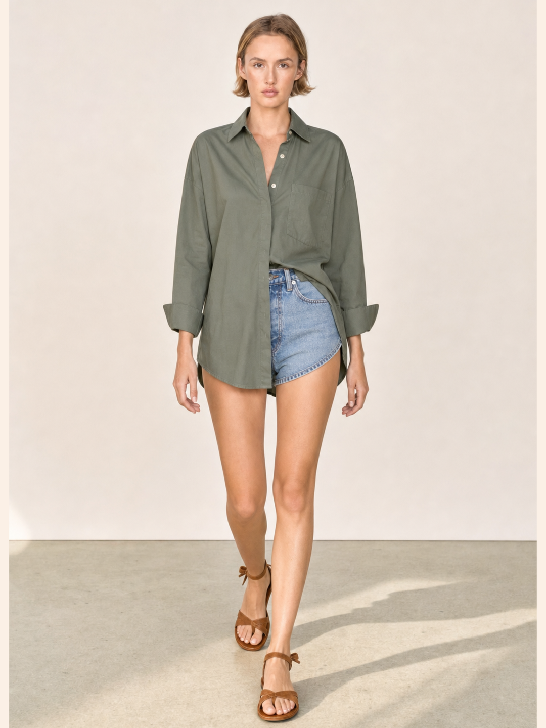 A woman modeling an oversized olive green button-down shirt with denim shorts and brown sandals, walking against a plain beige studio background.