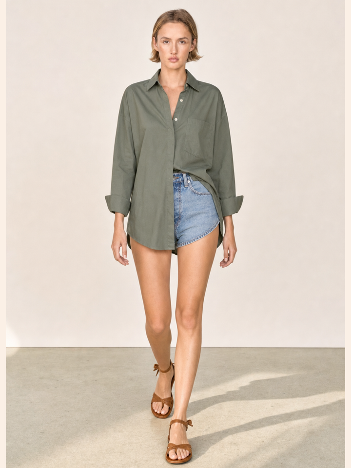 A woman modeling an oversized olive green button-down shirt with denim shorts and brown sandals, walking against a plain beige studio background.