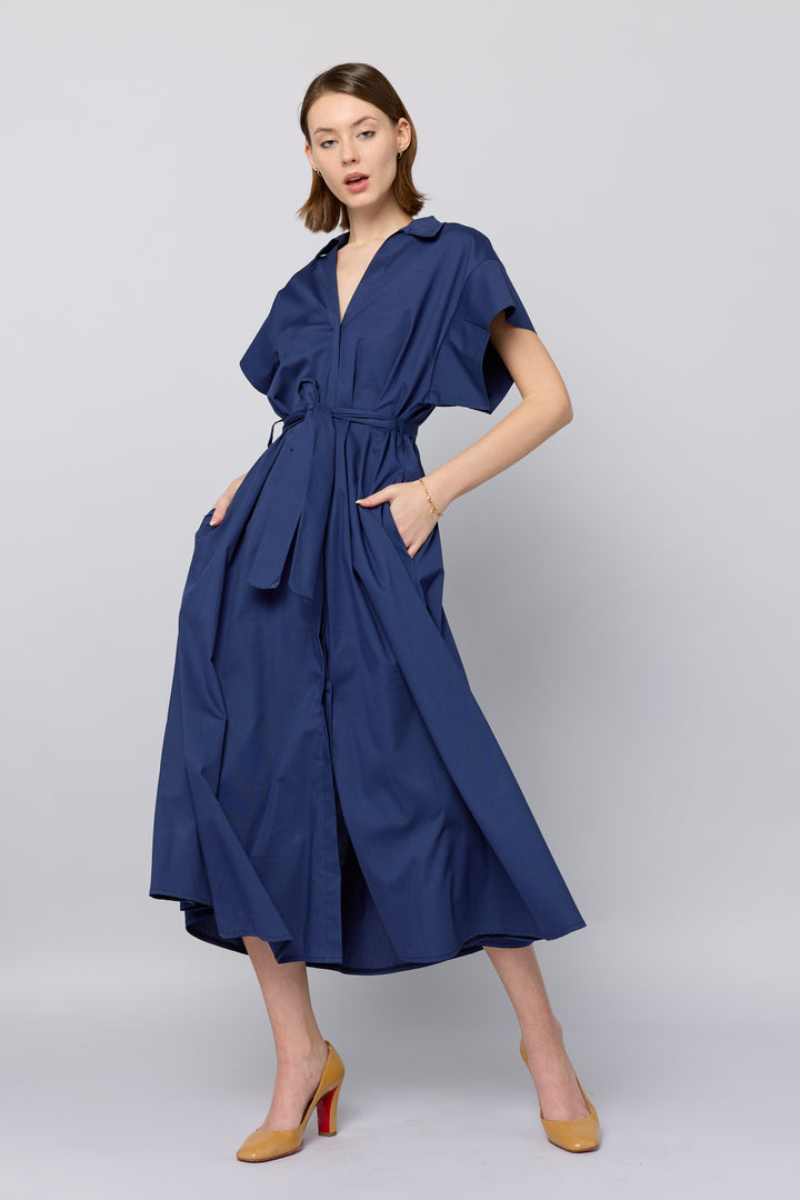 A woman models a midi-length shirt dress in pink with a classic collared neckline, short sleeves, a button-down front, and a matching fabric belt cinching the waist. The dress features side pockets and she is wearing open-toe heels.