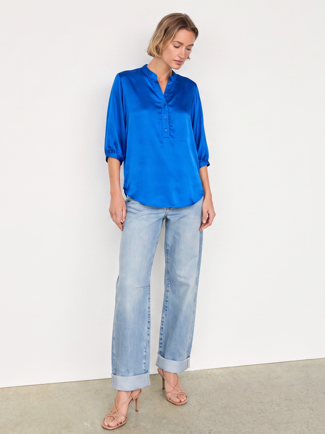 A woman modeling a bright royal blue satin blouse with three-quarter sleeves and a split neckline, paired with light-wash blue jeans and nude high-heeled sandals, standing against a plain white wall.