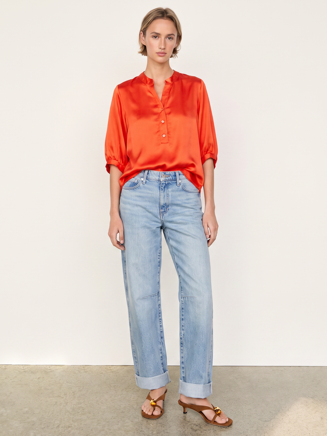 A model stands against a plain off-white wall wearing a bright orange satin button-up shirt, light-wash blue jeans, and brown heeled sandals.