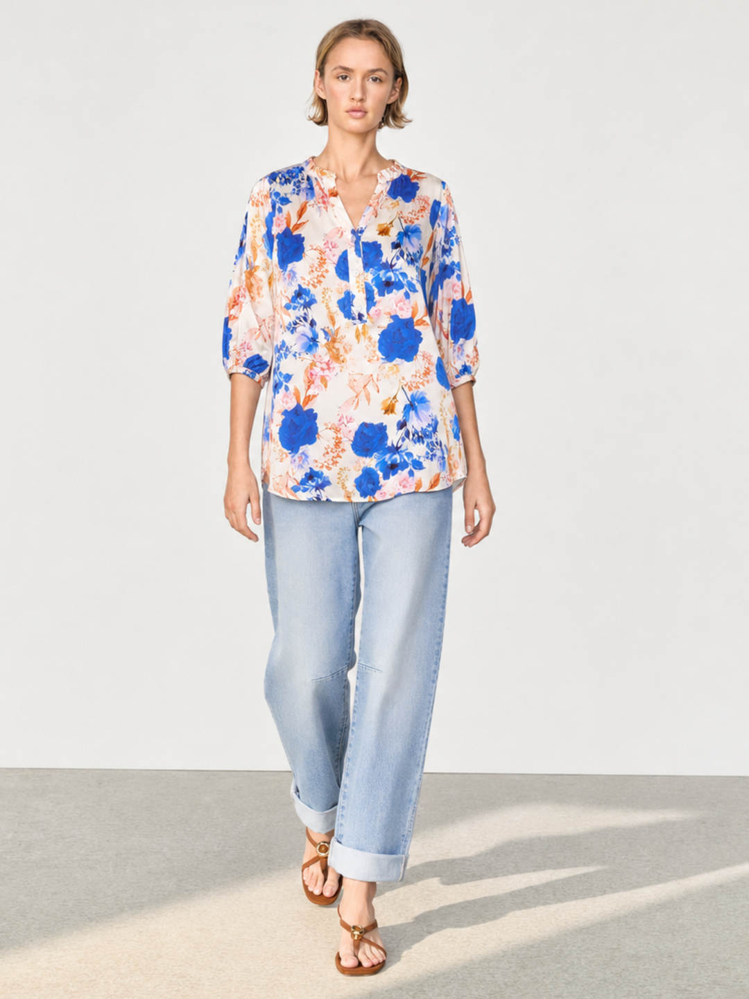 A woman modeling a short-sleeve button-up blouse with a blue, orange, and white abstract floral print, paired with light-wash cuffed jeans and brown sandals, set against a minimalist studio background.